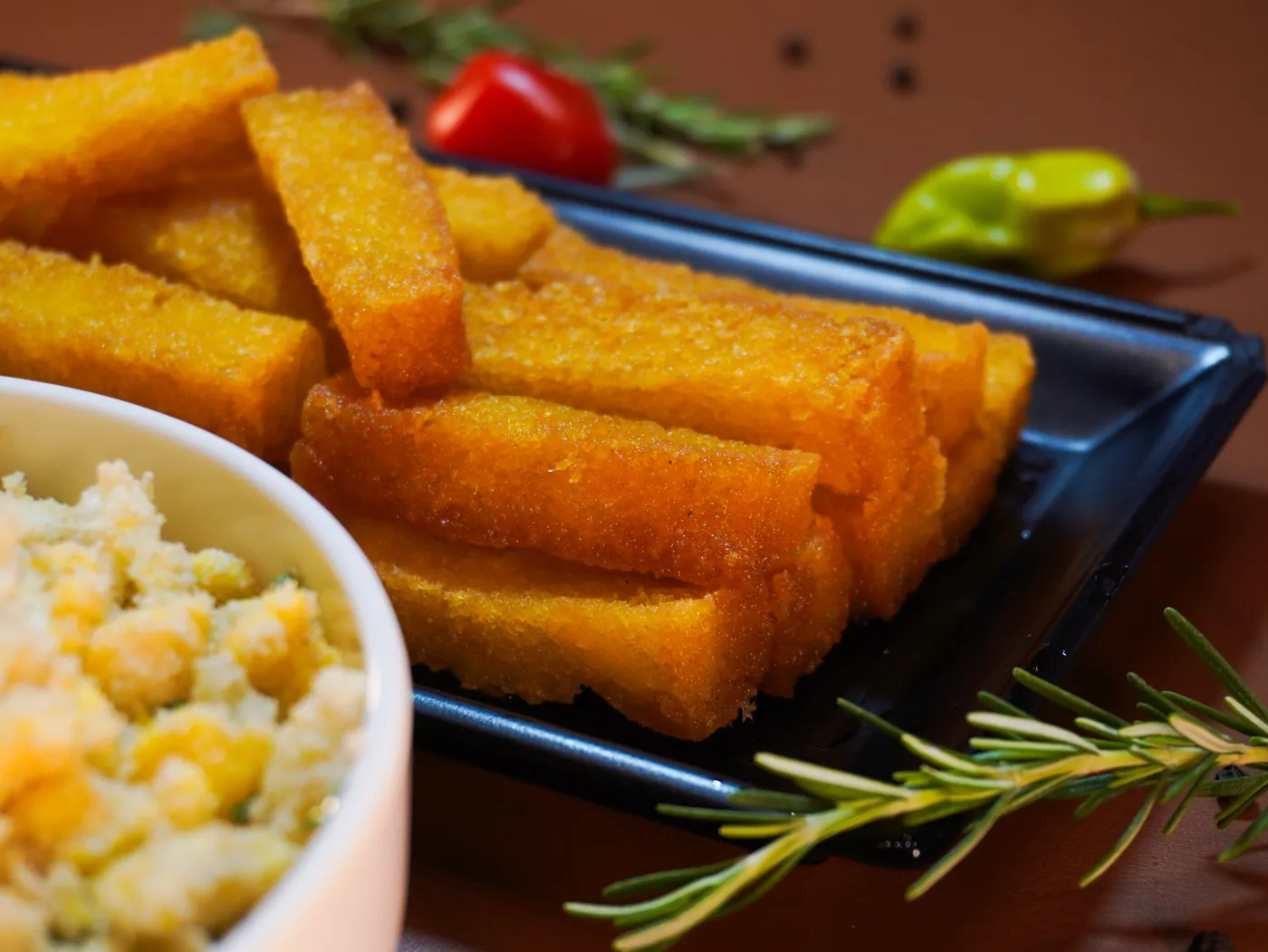 Polenta frita cortada grossa em travessa preta, com farofa de ovos ao lado
